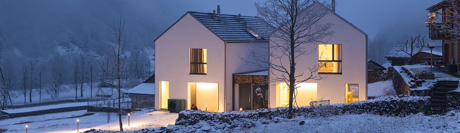 Upplýst hús í snjó með varmadælu úti við vegg